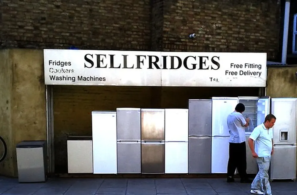 A used-appliance store lot with a row of secondhand refrigerators, one shopper checking an open fridge
